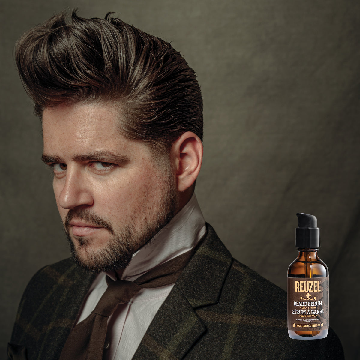 Male model with slicked up brown hair and beard in front of REUZEL beard serum