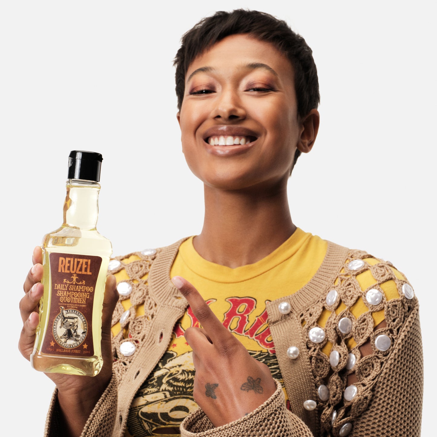 Female model with short black hair holding and pointing at REUZEL daily shampoo