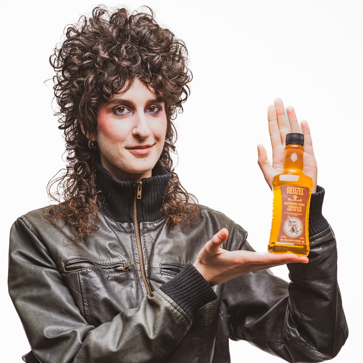 Female model with long brown curly hair holding REUZEL grooming tonic
