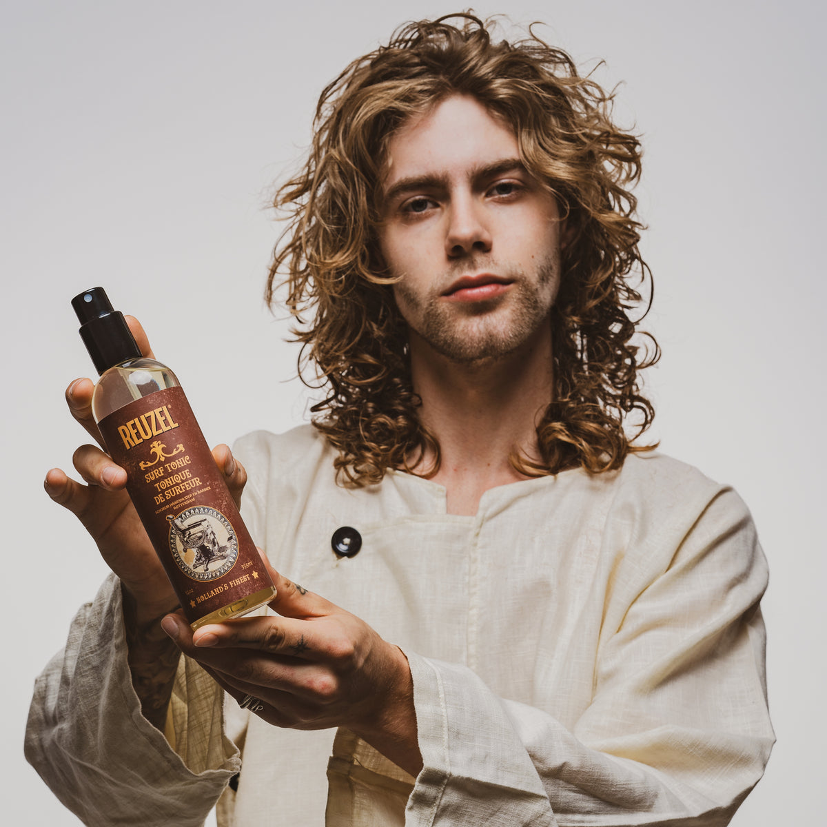 Male model with wavy brown hair holding REUZEL surf tonic sea salt spray