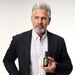 Male model with grey hair and beard holding REUZEL's Beard Oil