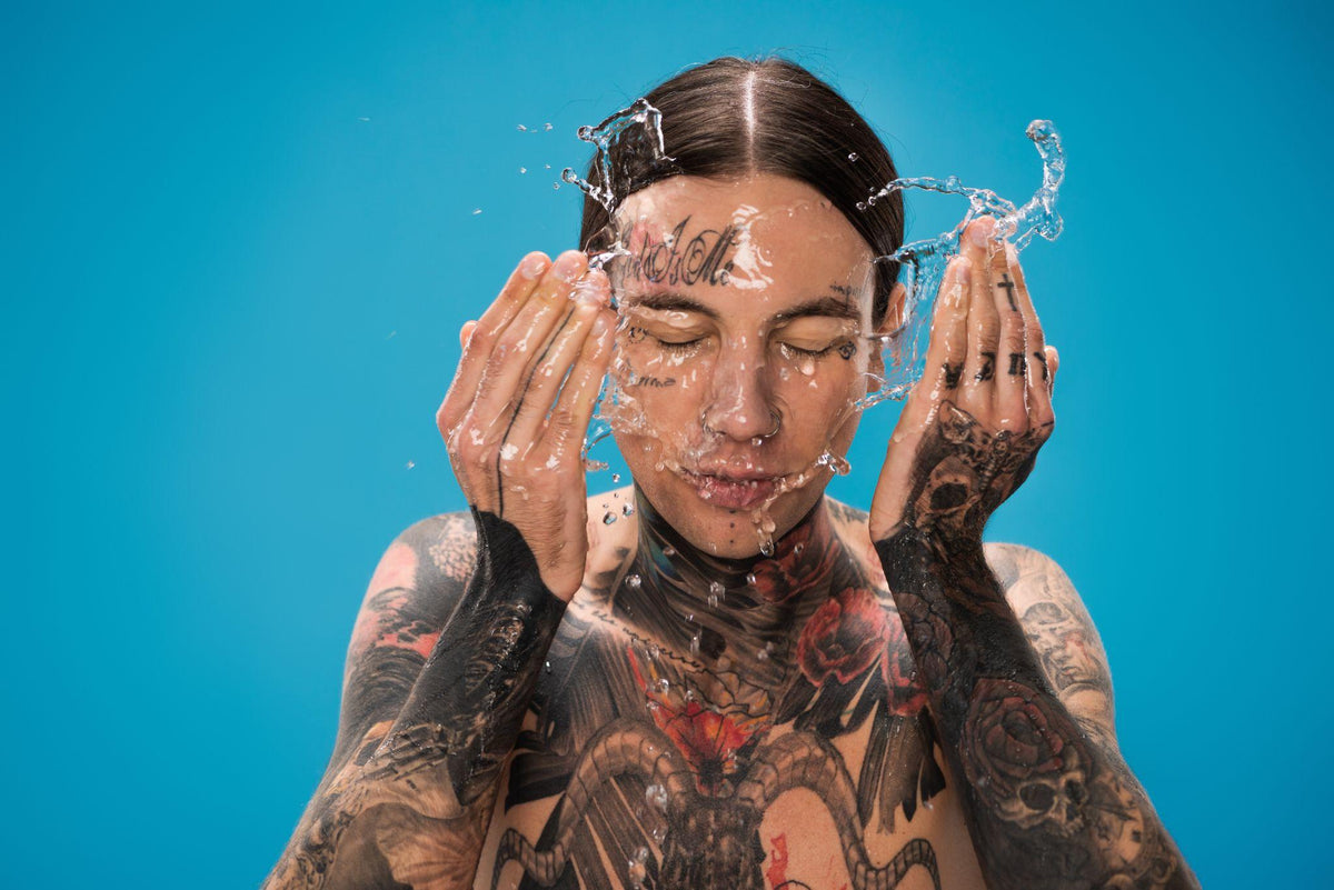 splash of water near young tattooed man washing face isolated on blue
