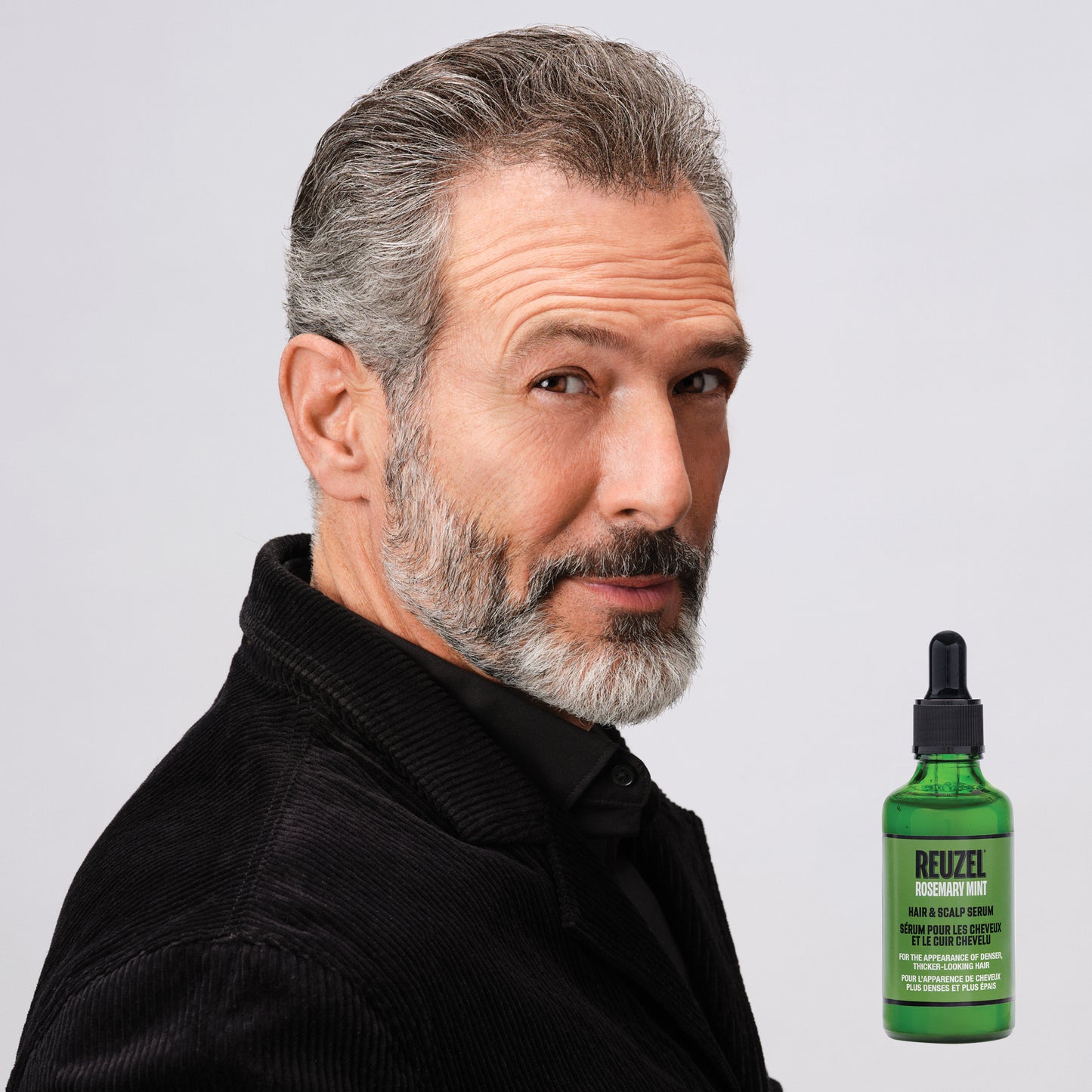 A man with gray hair and a gray beard wearing a black corduroy jacket, shown next to a green bottle of REUZEL rosemary mint Hair & Scalp Serum on a light gray background.