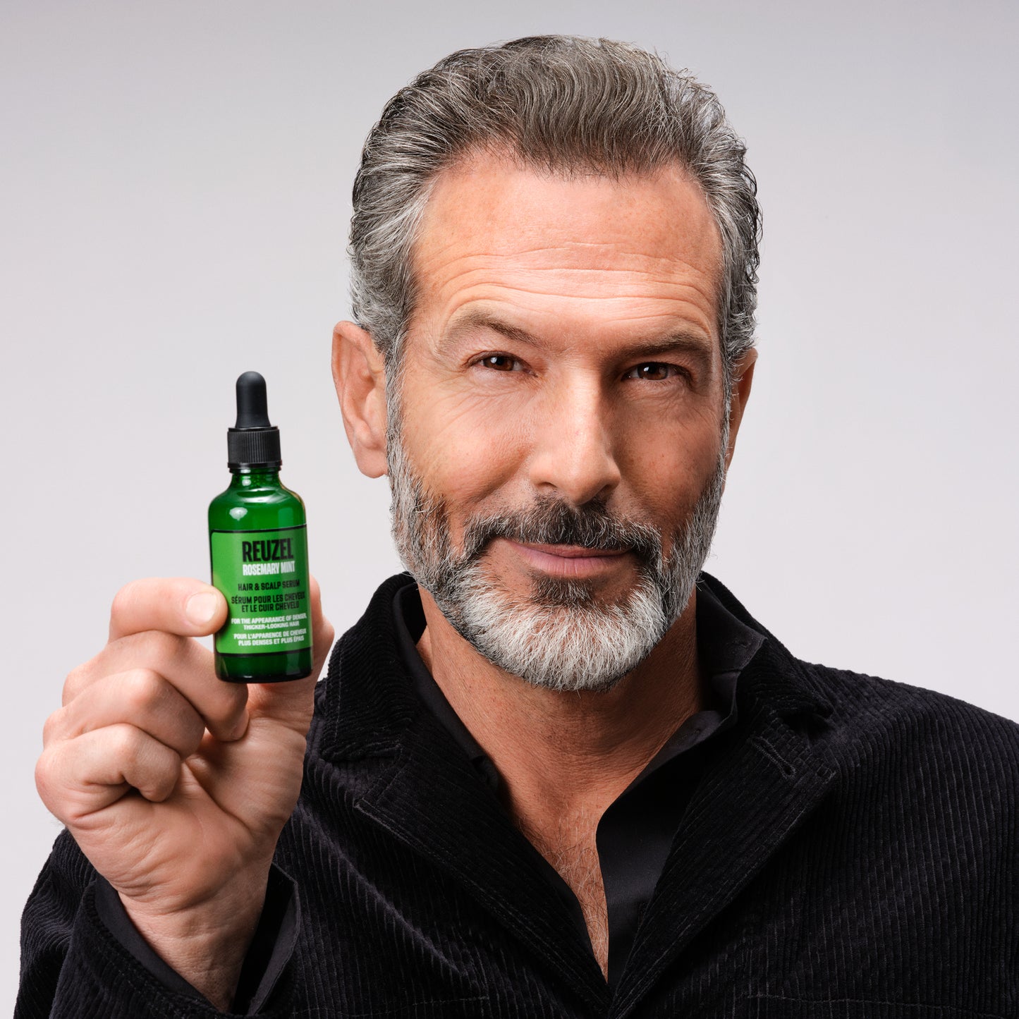 A man with gray hair and a graying beard holds up a small green dropper bottle of REUZEL rosemary mint hair and scalp serum