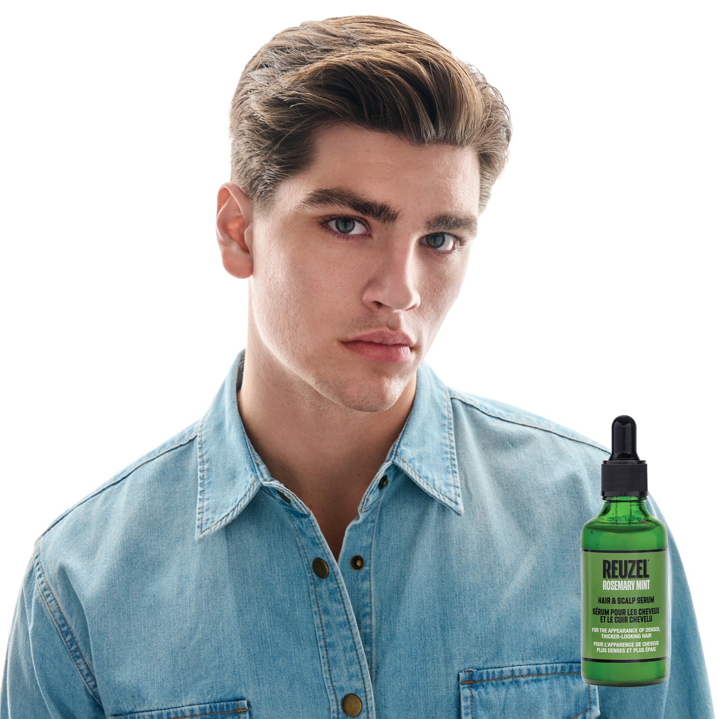 A young man with styled brown hair wearing a light blue denim shirt, shown against a white background with a green bottle of RUEZEL rosemary mint Hair & Scalp Serum positioned near his lower right.