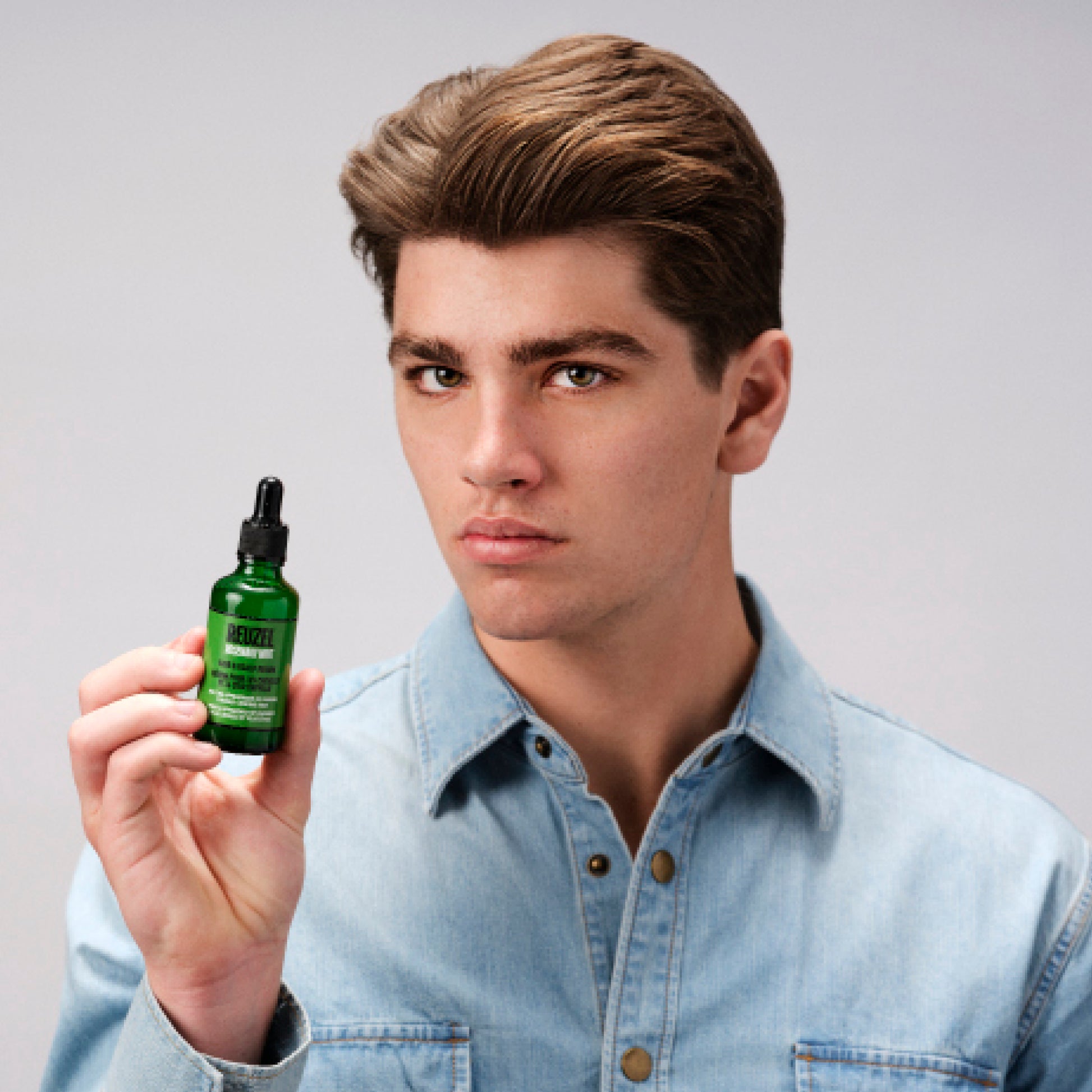 A young man with styled brown hair wearing a light blue denim shirt holds up a small green glass dropper bottle of REUZEL rosemary mint hair & scalp serum against a plain light gray studio background.