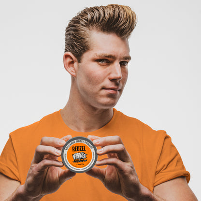 A man in an orange t-shirt holds a tin of REUZEL Orange Extreme Hold Matte Pomade in front of his chest, showing the product packaging against a light gray background.