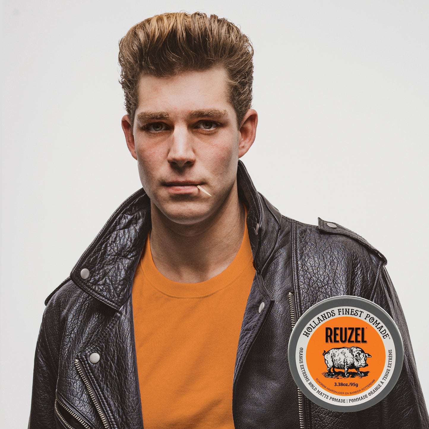 A young man with styled light brown hair wears a black leather jacket over an orange t-shirt, with REUZEL Orange Extreme Hold Matte Pomade. He is looking directly at the camera against a light gray background.