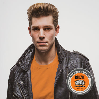 A young man with styled light brown hair wears a black leather jacket over an orange t-shirt, with REUZEL Orange Extreme Hold Matte Pomade. He is looking directly at the camera against a light gray background.