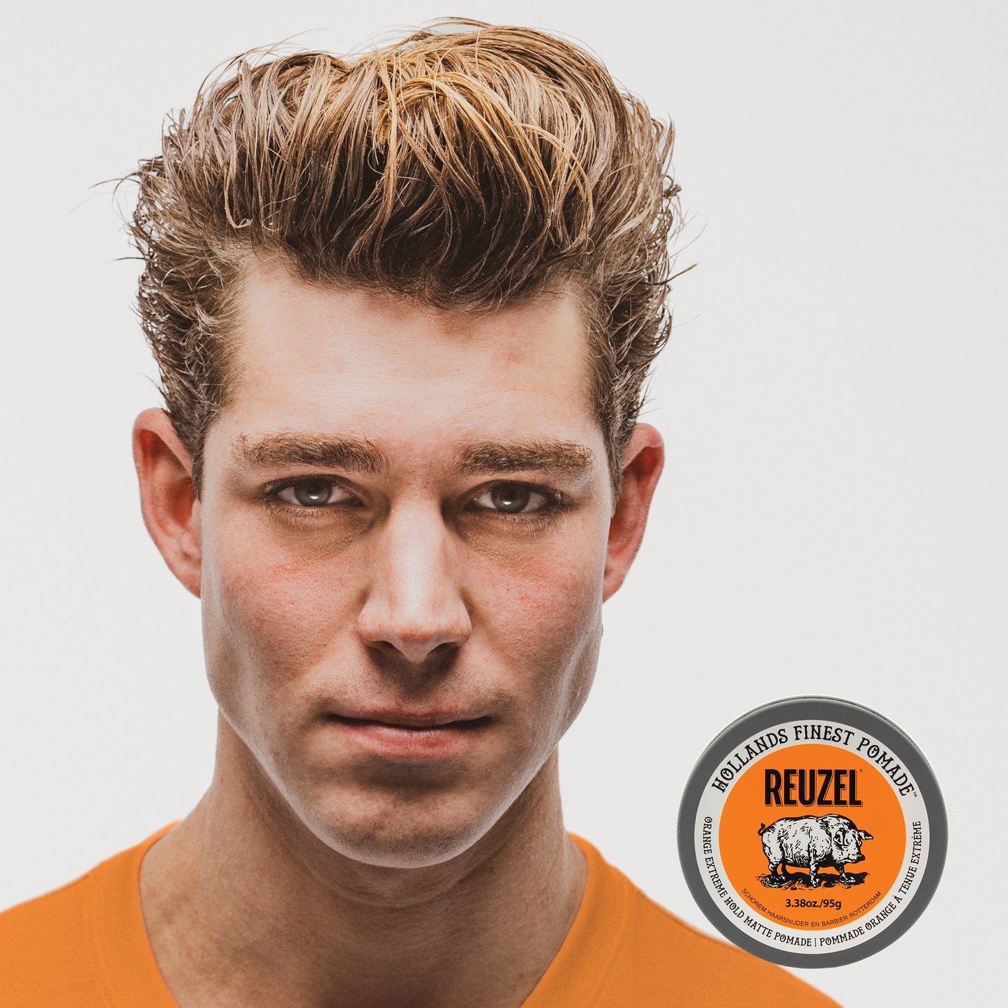 A close-up portrait of a man with styled light brown hair wearing an orange shirt, with REUZEL Orange Extreme Hold Matte Pomade in the lower right corner on a light gray background.