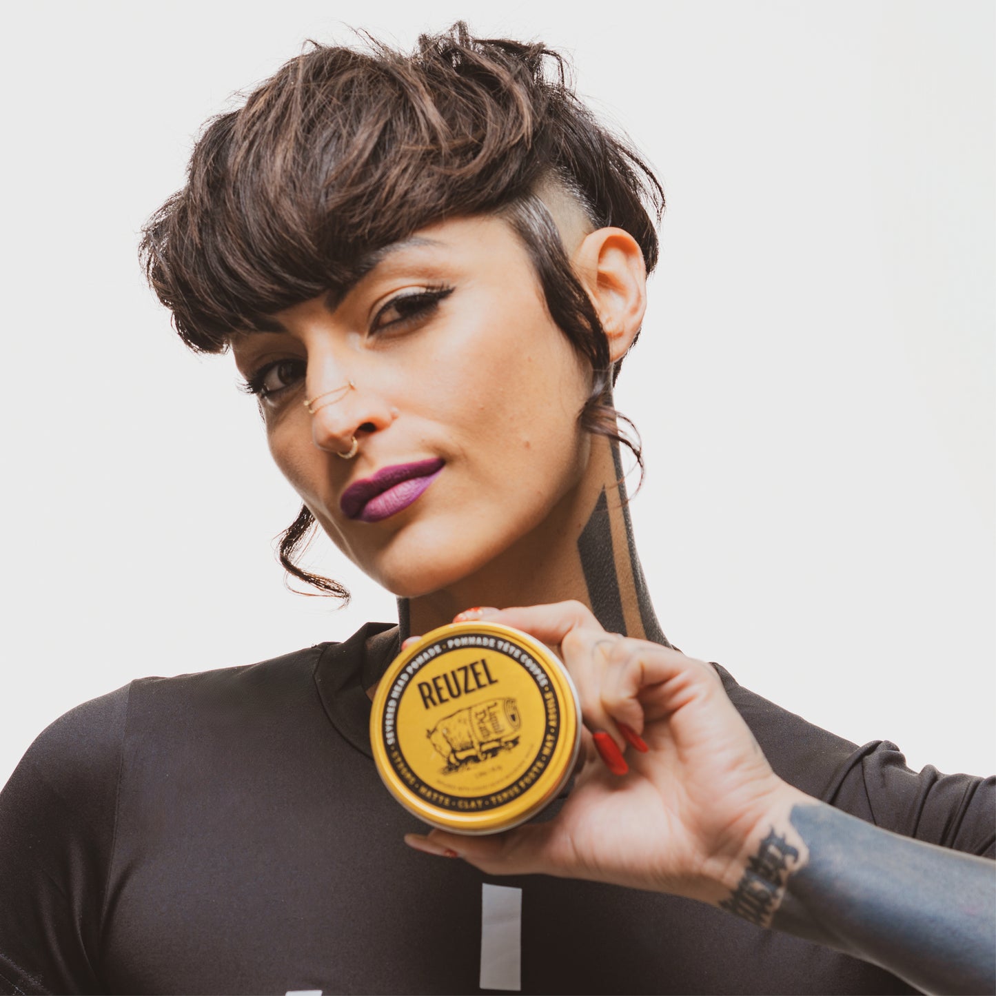 Woman with styled hair holding a puck of REUZEL x Liquid Death Severed Head Strong Matte Clay Pomad on a white background