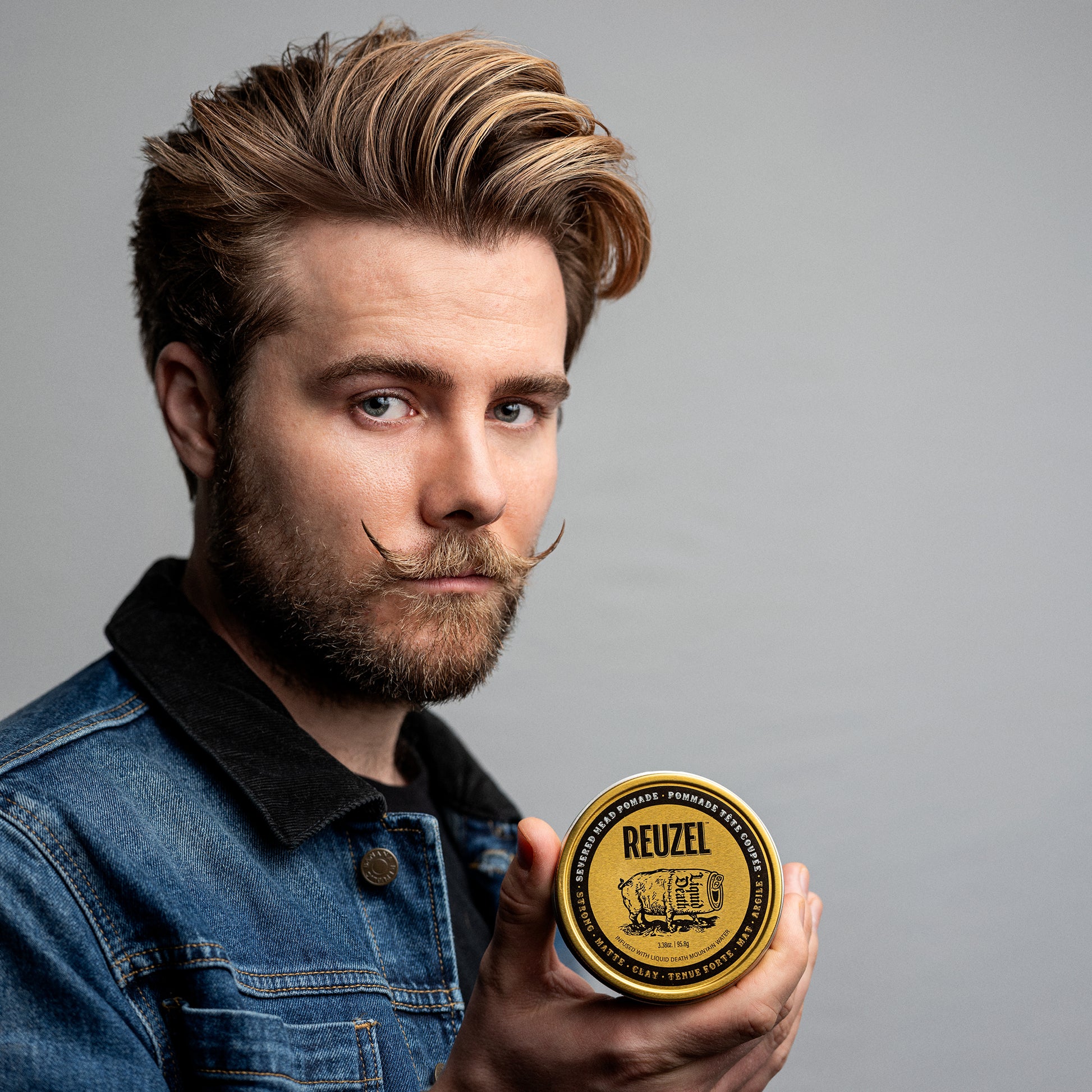 Male model with brown short hair and handlebar mustache next to REUZEL severed head strong matte clay pomade